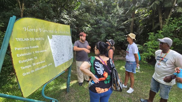 Foto em &aacute;rea vegetal de pessoal de p&eacute; ouvindo um guia, que est&aacute; ao lado de uma placa.