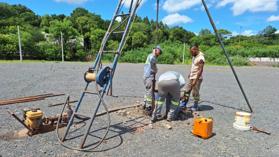 Trabalhadores realizam a&ccedil;&otilde;es de medi&ccedil;&atilde;o no terreno com britas. Ao fundo, um bosque verde e o c&eacute;u azul. 