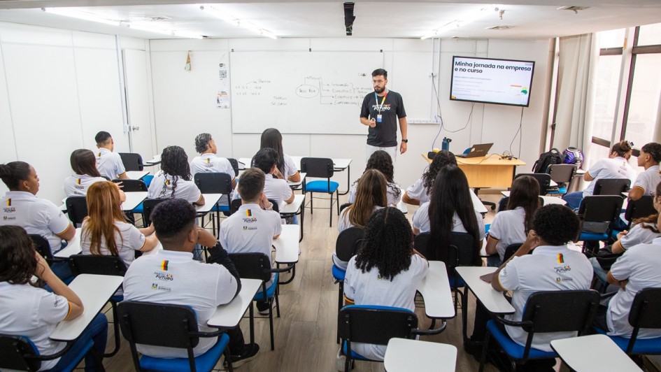 Programa Partiu Futuro Reconstru&ccedil;&atilde;o
A imagem retrata uma sala de aula moderna e cheia durante uma atividade de capacita&ccedil;&atilde;o ou aula te&oacute;rica.

Pessoas e Disposi&ccedil;&atilde;o:

Instrutor: No centro ao fundo, um homem jovem, de barba e &oacute;culos, veste uma camiseta preta com um logotipo colorido e cal&ccedil;as claras. Ele est&aacute; em p&eacute;, em frente &agrave; turma, em uma postura de fala e ensino.

Alunos: V&aacute;rios jovens (vistos de costas) preenchem a sala, sentados em cadeiras universit&aacute;rias pretas com assentos azuis e pranchetas brancas para escrita. Todos vestem uniformes padronizados: camisetas polo brancas com um logotipo colorido nas costas.

Elementos Pedag&oacute;gicos:

Quadro Branco: Na parede frontal, h&aacute; um quadro branco grande com anota&ccedil;&otilde;es manuais e diagramas sobre o curso.

Recursos Audiovisuais: &Agrave; direita, uma televis&atilde;o de tela plana fixada na parede exibe um slide de apresenta&ccedil;&atilde;o com o t&iacute;tulo "Minha jornada na empresa e no curso". Abaixo da TV, h&aacute; uma mesa de madeira clara com um notebook aberto e uma garrafa d'&aacute;gua.

Ambiente:

Estrutura: A sala &eacute; bem iluminada, com paredes brancas, piso laminado que imita madeira clara e teto branco com ilumina&ccedil;&atilde;o embutida.

Luz Natural: No lado direito da imagem, grandes janelas de vidro permitem a entrada de luz natural, sugerindo um ambiente arejado.