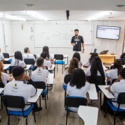 Programa Partiu Futuro Reconstru&ccedil;&atilde;o
A imagem retrata uma sala de aula moderna e cheia durante uma atividade de capacita&ccedil;&atilde;o ou aula te&oacute;rica.

Pessoas e Disposi&ccedil;&atilde;o:

Instrutor: No centro ao fundo, um homem jovem, de barba e &oacute;culos, veste uma camiseta preta com um logotipo colorido e cal&ccedil;as claras. Ele est&aacute; em p&eacute;, em frente &agrave; turma, em uma postura de fala e ensino.

Alunos: V&aacute;rios jovens (vistos de costas) preenchem a sala, sentados em cadeiras universit&aacute;rias pretas com assentos azuis e pranchetas brancas para escrita. Todos vestem uniformes padronizados: camisetas polo brancas com um logotipo colorido nas costas.

Elementos Pedag&oacute;gicos:

Quadro Branco: Na parede frontal, h&aacute; um quadro branco grande com anota&ccedil;&otilde;es manuais e diagramas sobre o curso.

Recursos Audiovisuais: &Agrave; direita, uma televis&atilde;o de tela plana fixada na parede exibe um slide de apresenta&ccedil;&atilde;o com o t&iacute;tulo "Minha jornada na empresa e no curso". Abaixo da TV, h&aacute; uma mesa de madeira clara com um notebook aberto e uma garrafa d'&aacute;gua.

Ambiente:

Estrutura: A sala &eacute; bem iluminada, com paredes brancas, piso laminado que imita madeira clara e teto branco com ilumina&ccedil;&atilde;o embutida.

Luz Natural: No lado direito da imagem, grandes janelas de vidro permitem a entrada de luz natural, sugerindo um ambiente arejado.