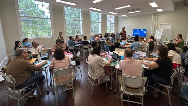 Foto mostra sala com coordenadores regionais em mesas circulares. Eles acompanham instru&ccedil;&otilde;es do palestrante. A sala tem janelas grandes que mostram &aacute;rvores. 