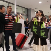 Recep&ccedil;&atilde;o Tur&iacute;stica no Rio Grande do Sul
A imagem retrata um momento de recep&ccedil;&atilde;o calorosa em um ambiente interno moderno e bem iluminado, possivelmente o sagu&atilde;o de um aeroporto ou centro de eventos.

Intera&ccedil;&atilde;o &agrave; Esquerda: Dois homens aparecem em primeiro plano, sorrindo e se cumprimentando. Um deles veste uma jaqueta preta e o outro uma camisa polo listrada em vermelho, azul e branco. Este &uacute;ltimo segura um smartphone e uma sacola preta com al&ccedil;as verdes.

Figura Central (Cultura Local): No centro da imagem, uma mulher vestida com um traje t&iacute;pico ga&uacute;cho (vestido longo marrom escuro com detalhes em renda e gola clara) segura duas sacolas promocionais pretas. As sacolas possuem al&ccedil;as verdes e a frase "SEJA BEM-VINDO AO RIO GRANDE DO SUL" estampada em letras verdes vibrantes.

Est&aacute;tua Viva: No canto direito, um artista perform&aacute;tico atua como uma est&aacute;tua viva. Ele est&aacute; inteiramente coberto por tinta cinza-chumbo, incluindo pele e roupas (camisa r&uacute;stica, cal&ccedil;as e uma faixa na cabe&ccedil;a), mantendo uma pose est&aacute;tica enquanto segura o que parece ser um chicote ou corda.

Cen&aacute;rio e Detalhes: O ambiente possui piso de granito claro, teto branco com ilumina&ccedil;&atilde;o embutida e grandes divis&oacute;rias de vidro ao fundo. Uma mala de viagem vermelha com rodas est&aacute; posicionada no ch&atilde;o entre os personagens principais, refor&ccedil;ando o contexto de viagem e turismo.