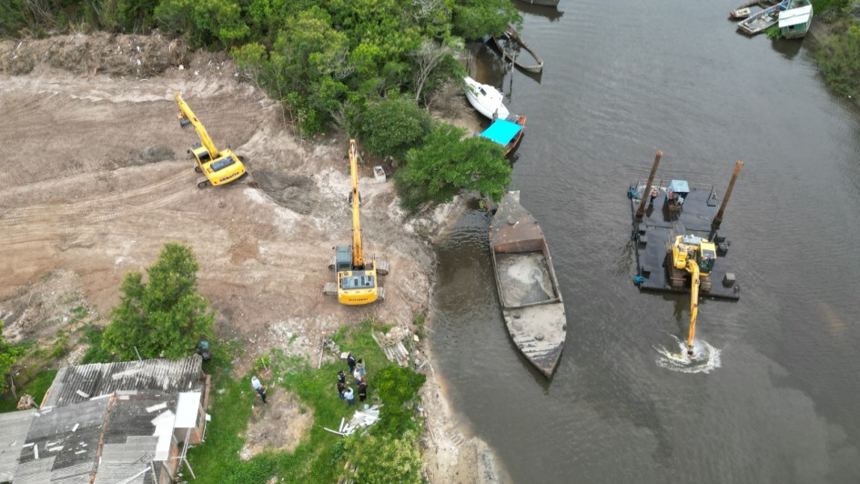Foto a&eacute;rea mostra rio com barco e draga em atividade e na margem com duas por&ccedil;&otilde;es de mata e no centro um terreno j&aacute; limpo por duas m&aacute;quinas retroescavadeiras. 