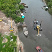 Foto a&eacute;rea mostra rio com barco e draga em atividade e na margem com duas por&ccedil;&otilde;es de mata e no centro um terreno j&aacute; limpo por duas m&aacute;quinas retroescavadeiras. 