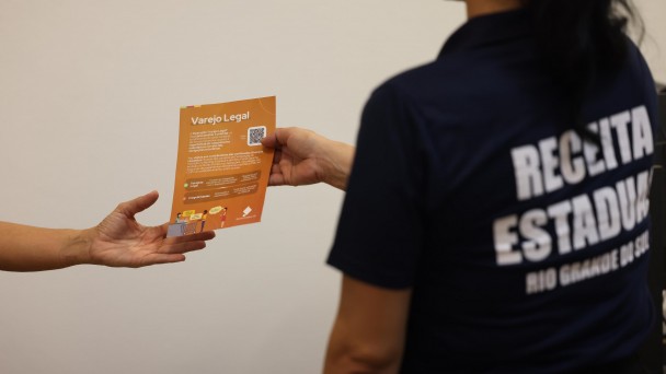 Uma pessoa de costas com camiseta da Receita Estadual entrega um panfleto a outra. Apenas a m&atilde;o aparece. No panfleto, &eacute; poss&iacute;vel ler "Varejo Legal". 