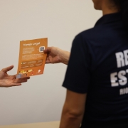 Uma pessoa de costas com camiseta da Receita Estadual entrega um panfleto a outra. Apenas a m&atilde;o aparece. No panfleto, &eacute; poss&iacute;vel ler "Varejo Legal". 