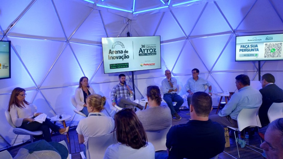 Esta &eacute; a descri&ccedil;&atilde;o detalhada da sexta imagem para fins de acessibilidade:

Imagem 6: Painel na Arena de Inova&ccedil;&atilde;o
A imagem registra uma palestra ou painel de discuss&atilde;o ocorrendo dentro de uma estrutura moderna durante um evento agr&iacute;cola.

Cen&aacute;rio e Ambiente:

A atividade acontece dentro de um domo geod&eacute;sico, com uma estrutura interna composta por tri&acirc;ngulos met&aacute;licos aparentes.

O ambiente &eacute; banhado por uma ilumina&ccedil;&atilde;o azulada tecnol&oacute;gica.

O piso &eacute; de deque de madeira clara.

Painelistas:

Cinco pessoas (duas mulheres e tr&ecirc;s homens) est&atilde;o sentadas em cadeiras brancas de design moderno, dispostas em semic&iacute;rculo.

Ao centro, um homem de camisa xadrez fala ao microfone. Os demais membros do painel ouvem atentamente ou consultam documentos.

Elementos Visuais e Informativos:

Tela Central: Um grande monitor exibe as marcas do evento: "Arena de Inova&ccedil;&atilde;o" e "36&ordf; Abertura Oficial da Colheita do Arroz". O texto na tela indica a data de 24 a 26 de fevereiro de 2026, em Embrapa - Cap&atilde;o do Le&atilde;o - RS.

Tela Lateral: &Agrave; direita, um monitor menor exibe a mensagem "FA&Ccedil;A SUA PERGUNTA" junto a um QR Code e um n&uacute;mero de WhatsApp para intera&ccedil;&atilde;o do p&uacute;blico.

P&uacute;blico: No primeiro plano, vemos as costas e cabe&ccedil;as de alguns espectadores sentados, focados na apresenta&ccedil;&atilde;o.