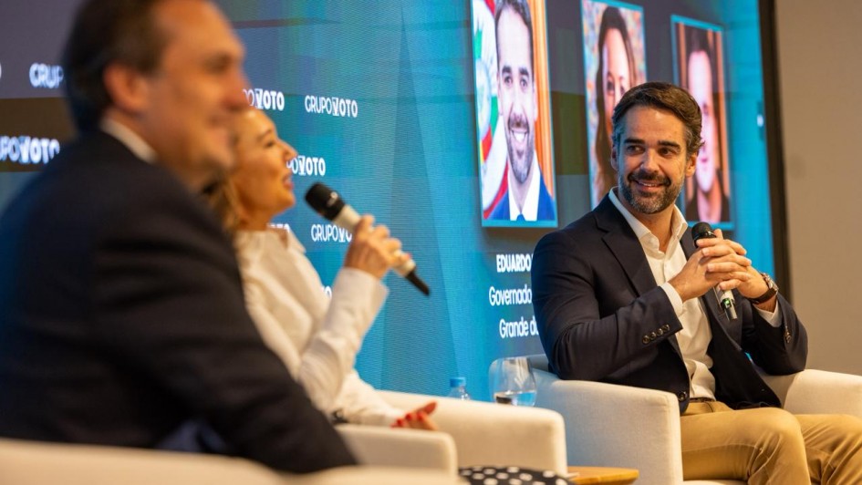 Em um palco de evento, tr&ecirc;s pessoas est&atilde;o sentadas em poltronas brancas participando de um painel. Leite, de terno escuro, segura um microfone e sorri enquanto olha para uma mulher ao lado, que tamb&eacute;m fala ao microfone. Ao fundo, um tel&atilde;o azul exibe fotos dos participantes e textos do evento.