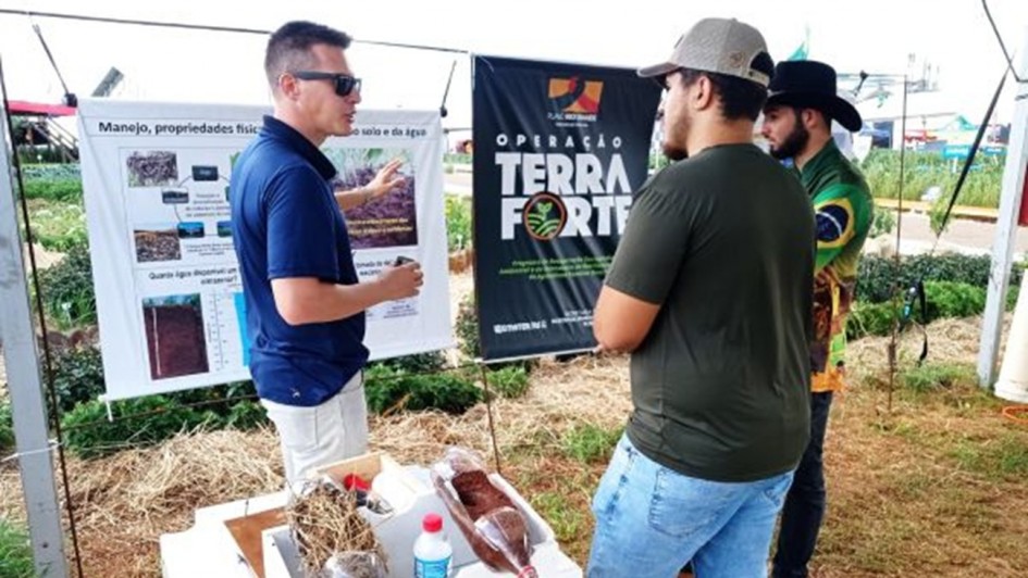 A fotografia retrata uma demonstra&ccedil;&atilde;o t&eacute;cnica durante um evento ao ar livre, identificado pelo nome do arquivo como a ExpoAgro Cotricampo. A cena foca na dissemina&ccedil;&atilde;o de conhecimento sobre manejo e conserva&ccedil;&atilde;o de recursos naturais.

Contexto e Pessoas
A&ccedil;&atilde;o Principal: Um homem &agrave; esquerda, vestindo uma camisa polo azul e &oacute;culos de sol, gesticula enquanto explica conte&uacute;dos t&eacute;cnicos para dois visitantes.

Visitantes: &Agrave; direita, dois homens escutam a explica&ccedil;&atilde;o. Um deles usa uma camiseta verde e bon&eacute; cinza; o outro veste uma camisa com estampas que remetem &agrave; bandeira do Brasil e um chap&eacute;u de aba larga preto.

Elementos de Demonstra&ccedil;&atilde;o
Cartazes T&eacute;cnicos: Atr&aacute;s do palestrante, h&aacute; um cartaz branco com o t&iacute;tulo "Manejo, propriedades f&iacute;sicas [do] solo e da &aacute;gua", contendo diagramas e fotos explicativas. Ao lado, um banner vertical preto exibe o logotipo da "Opera&ccedil;&atilde;o Terra Forte".

Experimento Pr&aacute;tico: Em primeiro plano, sobre uma bancada branca, h&aacute; uma demonstra&ccedil;&atilde;o did&aacute;tica utilizando garrafas pl&aacute;sticas cortadas. As garrafas cont&ecirc;m diferentes camadas de solo e cobertura vegetal (palha), provavelmente utilizadas para mostrar visualmente processos de infiltra&ccedil;&atilde;o de &aacute;gua e prote&ccedil;&atilde;o do solo contra eros&atilde;o.

Ambiente
Cen&aacute;rio: O evento ocorre em uma &aacute;rea aberta com gramado e canteiros de plantas ao fundo. Estruturas de metal e lonas sugerem a montagem de estandes t&iacute;picos de feiras agr&iacute;colas.