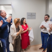A imagem mostra um grupo de pessoas em p&eacute;, batendo palmas e sorrindo, em frente a uma placa de inaugura&ccedil;&atilde;o afixada na parede branca de um hospital. 