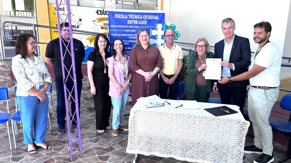 A fotografia registra um ato oficial realizado em uma escola, identificado pelo t&iacute;tulo do arquivo como a autoriza&ccedil;&atilde;o para o in&iacute;cio de obras na Escola T&eacute;cnica Estadual Entre-Iju&iacute;s.

Contexto e Pessoas
A&ccedil;&atilde;o Principal: &Agrave; direita da imagem, um homem em um blazer escuro e uma mulher idosa seguram juntos um documento oficial, exibindo-o para a c&acirc;mera. Um homem mais jovem, &agrave; extrema direita, tamb&eacute;m toca o documento, sinalizando um ato de assinatura ou entrega formal.

Grupo: Ao todo, nove pessoas (cinco mulheres e quatro homens) est&atilde;o posicionadas em p&eacute; atr&aacute;s de uma mesa, posando para a foto com express&otilde;es formais e sorridentes.

Elementos de Identifica&ccedil;&atilde;o
Banner Informativo: No centro, ao fundo, h&aacute; um banner azul com o nome "Escola T&eacute;cnica Estadual Entre-Iju&iacute;s". O banner lista n&iacute;veis de ensino e cursos, como Ensino Fundamental, Ensino M&eacute;dio e T&eacute;cnico em Administra&ccedil;&atilde;o.

Mesa Cerimonial: &Agrave; frente do grupo, h&aacute; uma mesa coberta com uma toalha de renda branca. Sobre ela, repousam uma pasta, uma caneta e um documento, refor&ccedil;ando o contexto de uma assinatura oficial.

Ambiente
Cen&aacute;rio Escolar: O evento ocorre em uma &aacute;rea coberta da escola. A parede ao fundo possui desenhos coloridos e palavras de incentivo como "Educa&ccedil;&atilde;o" e "Transforma&ccedil;&atilde;o".

Detalhes Estruturais: O piso &eacute; de pedras irregulares em tons de marrom e cinza. &Agrave; esquerda, nota-se uma estrutura met&aacute;lica (treli&ccedil;a) pintada de roxo e algumas cadeiras azuis empilhadas ao fundo.