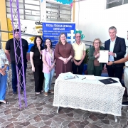 A fotografia registra um ato oficial realizado em uma escola, identificado pelo t&iacute;tulo do arquivo como a autoriza&ccedil;&atilde;o para o in&iacute;cio de obras na Escola T&eacute;cnica Estadual Entre-Iju&iacute;s.

Contexto e Pessoas
A&ccedil;&atilde;o Principal: &Agrave; direita da imagem, um homem em um blazer escuro e uma mulher idosa seguram juntos um documento oficial, exibindo-o para a c&acirc;mera. Um homem mais jovem, &agrave; extrema direita, tamb&eacute;m toca o documento, sinalizando um ato de assinatura ou entrega formal.

Grupo: Ao todo, nove pessoas (cinco mulheres e quatro homens) est&atilde;o posicionadas em p&eacute; atr&aacute;s de uma mesa, posando para a foto com express&otilde;es formais e sorridentes.

Elementos de Identifica&ccedil;&atilde;o
Banner Informativo: No centro, ao fundo, h&aacute; um banner azul com o nome "Escola T&eacute;cnica Estadual Entre-Iju&iacute;s". O banner lista n&iacute;veis de ensino e cursos, como Ensino Fundamental, Ensino M&eacute;dio e T&eacute;cnico em Administra&ccedil;&atilde;o.

Mesa Cerimonial: &Agrave; frente do grupo, h&aacute; uma mesa coberta com uma toalha de renda branca. Sobre ela, repousam uma pasta, uma caneta e um documento, refor&ccedil;ando o contexto de uma assinatura oficial.

Ambiente
Cen&aacute;rio Escolar: O evento ocorre em uma &aacute;rea coberta da escola. A parede ao fundo possui desenhos coloridos e palavras de incentivo como "Educa&ccedil;&atilde;o" e "Transforma&ccedil;&atilde;o".

Detalhes Estruturais: O piso &eacute; de pedras irregulares em tons de marrom e cinza. &Agrave; esquerda, nota-se uma estrutura met&aacute;lica (treli&ccedil;a) pintada de roxo e algumas cadeiras azuis empilhadas ao fundo.