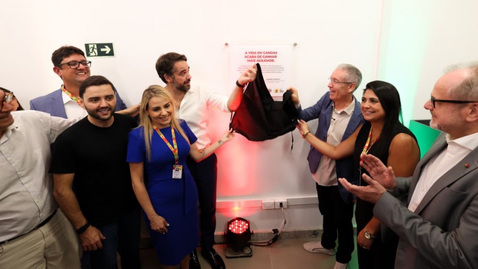 Um grupo de autoridades, incluindo o governador Eduardo Leite e o vice-governador Gabriel Souza, est&aacute; reunido para o descerramento de uma placa inaugural na parede branca. O governador segura o tecido preto que cobria a placa, revelando um texto que come&ccedil;a com a frase em destaque: "A vida em Canoas acaba de ganhar mais agilidade". As pessoas ao redor sorriem e aplaudem o momento.