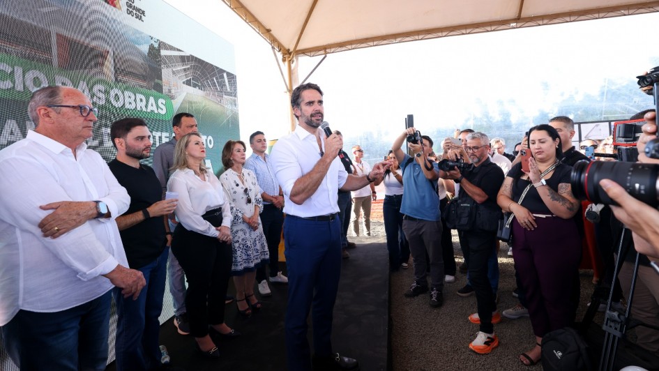 Eduardo Leite fala ao microfone em um palanque sob uma tenda branca. Ele veste camisa branca e cal&ccedil;a azul. Ao seu lado, autoridades acompanham o discurso em frente a um painel com a logomarca do Governo do Rio Grande do Sul e proje&ccedil;&otilde;es da futura escola. &Agrave; direita, fot&oacute;grafos e cinegrafistas registram o evento.