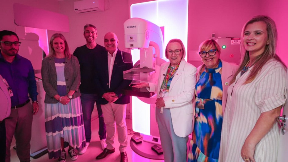 Esta &eacute; a descri&ccedil;&atilde;o detalhada da oitava imagem para fins de acessibilidade:

Imagem 8: Entrega de Mam&oacute;grafo na Santa Casa de Pelotas
A imagem retrata a cerim&ocirc;nia de entrega ou inaugura&ccedil;&atilde;o de um novo equipamento m&eacute;dico em uma sala de exames hospitalares.

Equipamento Central: No centro da imagem, destaca-se um mam&oacute;grafo moderno da marca Siemens. O aparelho &eacute; branco e possui um grande painel retangular ao fundo que emite uma luz branca intensa, iluminando a cena.

Pessoas: Um grupo de sete pessoas (quatro mulheres e tr&ecirc;s homens) est&aacute; posicionado ao redor do equipamento.

Todos est&atilde;o sorrindo para a c&acirc;mera em uma postura formal.

Uma mulher ao centro, vestindo um blazer branco e cal&ccedil;as azuis claras, estende a m&atilde;o em dire&ccedil;&atilde;o ao mam&oacute;grafo, apresentando-o.

Os demais vestem roupas casuais e sociais, incluindo blazers, vestidos coloridos e camisas polo.

Ambiente e Ilumina&ccedil;&atilde;o:

A sala &eacute; banhada por uma ilumina&ccedil;&atilde;o ambiente em tons de rosa e magenta, possivelmente em alus&atilde;o &agrave; campanha de conscientiza&ccedil;&atilde;o do Outubro Rosa.

As paredes s&atilde;o de cor clara e o teto possui dois aparelhos de ar-condicionado instalados.

Ao fundo, &agrave; direita, h&aacute; um painel el&eacute;trico com uma luz verde acesa e a indica&ccedil;&atilde;o de voltagem "220".

Clima: A imagem transmite uma sensa&ccedil;&atilde;o de conquista, celebra&ccedil;&atilde;o e avan&ccedil;o na infraestrutura de sa&uacute;de local.