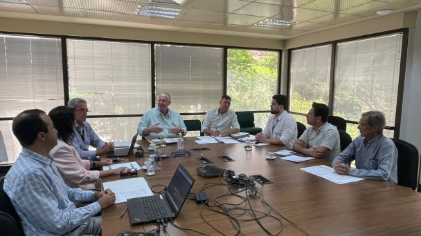 Foto de uma sala de reuni&otilde;es, com uma grande mesa marrom e janelas grandes ao fundo. H&aacute; sete homens e uma mulher sentados ao redor da mesa. A maior parte olha para um homem idoso que gesticula enquanto fala.