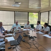 Foto de uma sala de reuni&otilde;es, com uma grande mesa marrom e janelas grandes ao fundo. H&aacute; sete homens e uma mulher sentados ao redor da mesa. A maior parte olha para um homem idoso que gesticula enquanto fala.