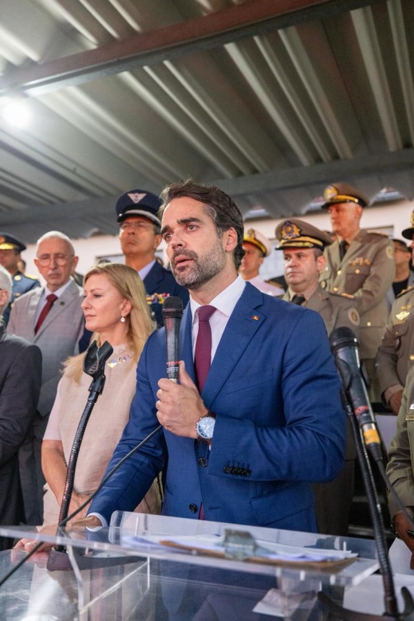 As imagens retratam uma cerimônia oficial de posse e anúncio de investimentos no Rio Grande do Sul. Abaixo, as descrições detalhadas para fins de acessibilidade: Imagem 1: Posse do Comando-Geral da Brigada Militar Esta imagem apresenta um plano médio lateral do governador Eduardo Leite durante seu discurso. O Governador: Localizado à direita da foto, Eduardo Leite está posicionado atrás de um púlpito de acrílico transparente. Ele veste um paletó azul, camisa branca e gravata bordô, segurando o microfone com as duas mãos enquanto fala. Autoridades e Convidados: Atrás do governador, uma fileira de autoridades acompanha o ato. Entre elas, destacam-se diversos oficiais militares em uniformes formais (verde-oliva, caqui e azul-escuro) com medalhas e quepe. Há também civis em trajes sociais, incluindo uma mulher de blusa bege e um homem de terno cinza com gravata vermelha. Cenário: O evento ocorre sob uma estrutura de teto metálico aparente, com iluminação natural. Imagem 2: Destaque aos Investimentos em Estrutura Esta imagem é um plano mais fechado (close-up) da mesma cena anterior, focando na expressão do governador. Expressão: Eduardo Leite é mostrado em um ângulo ligeiramente inferior, olhando para frente com uma expressão séria e compenetrada enquanto discursa. Segundo Plano: A profundidade de campo é menor, deixando as autoridades ao fundo um pouco menos nítidas, mas ainda identificáveis, reforçando o foco na fala do governador sobre os investimentos realizados. Detalhes Técnicos: O microfone preto e a borda do púlpito de acrílico aparecem na parte inferior da moldura.