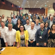 Foto posada num audit&oacute;rio. &Agrave; frente, h&aacute; uma mesa. Atr&aacute;s dela, at&eacute; o fundo do audit&oacute;rio aparecem mais de 50 pessoas de p&eacute;, que olham para a c&acirc;mera. Boa parte sorri.