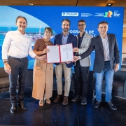 A imagem registra o momento da assinatura de um protocolo de inten&ccedil;&otilde;es para um investimento de R$ 130 milh&otilde;es no Distrito Industrial do Rio Grande.

Composi&ccedil;&atilde;o e Participantes
O Governador: No centro da foto, o governador Eduardo Leite segura uma pasta vermelha aberta contendo o documento assinado. Ele veste um blazer azul-marinho, camisa social azul-clara e cal&ccedil;a bege.

Autoridades: Ao lado do governador, quatro outras autoridades (tr&ecirc;s homens e uma mulher) tamb&eacute;m seguram as bordas da pasta, simbolizando a coopera&ccedil;&atilde;o entre as partes. Eles vestem trajes casuais-sociais, como blazers sem gravata e camisas claras.

Cen&aacute;rio: O grupo est&aacute; posicionado sobre um tablado preto em uma sala de arquitetura moderna, com forro e pain&eacute;is de madeira.

Elementos Visuais de Fundo
Painel Digital: Atr&aacute;s do grupo, um grande tel&atilde;o azul exibe as logomarcas da Portos RS, da Secretaria de Desenvolvimento Econ&ocirc;mico, da Secretaria de Log&iacute;stica e Transportes e o bras&atilde;o do Governo do Estado do Rio Grande do Sul.

Identidade Visual: O tel&atilde;o tamb&eacute;m apresenta uma fotografia a&eacute;rea do complexo portu&aacute;rio e industrial, refor&ccedil;ando o tema do investimento anunciado.