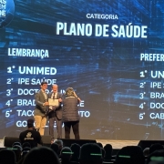 Foto mostra duas pessoas no palco recebendo o certificado com a lista de planos de sa&uacute;de citados na pesquisa listados no tel&atilde;o ao fundo.