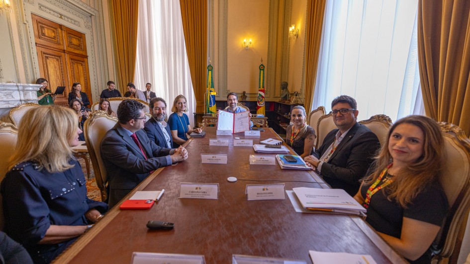A fotografia registra o momento em que o Governador Eduardo Leite apresenta o documento oficial assinado durante a reuni&atilde;o no Pal&aacute;cio Piratini.

A&ccedil;&atilde;o Central: No topo da longa mesa de madeira, o Governador Eduardo Leite segura, com as duas m&atilde;os, uma pasta vermelha aberta, exibindo um documento com o bras&atilde;o do Estado. Ele olha para a c&acirc;mera com um sorriso, simbolizando a oficializa&ccedil;&atilde;o do ato.

Os Participantes:

Sete pessoas est&atilde;o sentadas ao longo das laterais da mesa, voltadas em dire&ccedil;&atilde;o ao Governador ou &agrave; c&acirc;mera.

As mulheres e homens presentes vestem trajes executivos. Algumas das participantes &agrave; direita da imagem olham diretamente para o fot&oacute;grafo.

Detalhes da Mesa:

Sobre a mesa, h&aacute; diversas placas de identifica&ccedil;&atilde;o brancas. &Eacute; poss&iacute;vel ler nomes como "Juliana Debaquer", "Carolina Scarparo", "Regiane Lopes" e "Felipe Cristov&atilde;o".

H&aacute; tamb&eacute;m pastas de documentos, tablets e smartphones espalhados pela superf&iacute;cie de couro da mesa.

O Ambiente:

O sal&atilde;o possui p&eacute;-direito alto com paredes decoradas em tons de creme e molduras cl&aacute;ssicas.

Ao fundo, entre grandes janelas com cortinas amarelas/douradas, est&atilde;o posicionadas as bandeiras do Brasil e do Rio Grande do Sul.

&Agrave; esquerda, v&ecirc;-se a porta de madeira entalhada e parte de uma lareira de m&aacute;rmore.

Composi&ccedil;&atilde;o: A foto foi tirada da extremidade oposta da mesa, criando uma perspectiva de profundidade que conduz o olhar do espectador diretamente para o Governador e o documento no centro do fundo.
