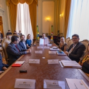 A fotografia registra o momento em que o Governador Eduardo Leite apresenta o documento oficial assinado durante a reuni&atilde;o no Pal&aacute;cio Piratini.

A&ccedil;&atilde;o Central: No topo da longa mesa de madeira, o Governador Eduardo Leite segura, com as duas m&atilde;os, uma pasta vermelha aberta, exibindo um documento com o bras&atilde;o do Estado. Ele olha para a c&acirc;mera com um sorriso, simbolizando a oficializa&ccedil;&atilde;o do ato.

Os Participantes:

Sete pessoas est&atilde;o sentadas ao longo das laterais da mesa, voltadas em dire&ccedil;&atilde;o ao Governador ou &agrave; c&acirc;mera.

As mulheres e homens presentes vestem trajes executivos. Algumas das participantes &agrave; direita da imagem olham diretamente para o fot&oacute;grafo.

Detalhes da Mesa:

Sobre a mesa, h&aacute; diversas placas de identifica&ccedil;&atilde;o brancas. &Eacute; poss&iacute;vel ler nomes como "Juliana Debaquer", "Carolina Scarparo", "Regiane Lopes" e "Felipe Cristov&atilde;o".

H&aacute; tamb&eacute;m pastas de documentos, tablets e smartphones espalhados pela superf&iacute;cie de couro da mesa.

O Ambiente:

O sal&atilde;o possui p&eacute;-direito alto com paredes decoradas em tons de creme e molduras cl&aacute;ssicas.

Ao fundo, entre grandes janelas com cortinas amarelas/douradas, est&atilde;o posicionadas as bandeiras do Brasil e do Rio Grande do Sul.

&Agrave; esquerda, v&ecirc;-se a porta de madeira entalhada e parte de uma lareira de m&aacute;rmore.

Composi&ccedil;&atilde;o: A foto foi tirada da extremidade oposta da mesa, criando uma perspectiva de profundidade que conduz o olhar do espectador diretamente para o Governador e o documento no centro do fundo.