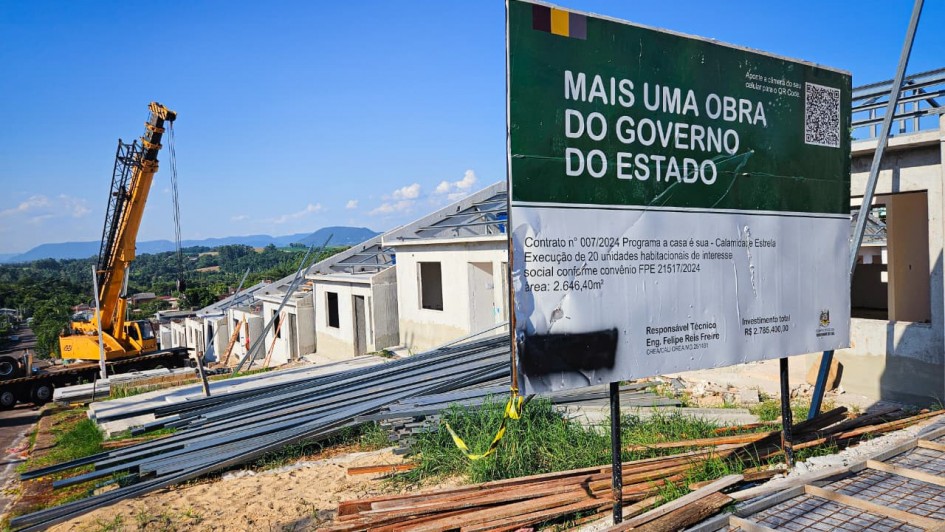 Esta &eacute; uma descri&ccedil;&atilde;o detalhada da imagem para fins de acessibilidade:

Descri&ccedil;&atilde;o da Imagem
A fotografia registra um canteiro de obras de um conjunto habitacional em est&aacute;gio intermedi&aacute;rio de constru&ccedil;&atilde;o, sob um c&eacute;u azul sem nuvens.

Placa Informativa (Primeiro Plano): No lado direito da imagem, h&aacute; uma grande placa de metal com as cores do estado (verde, vermelho e amarelo no topo).

O texto em destaque diz: "MAIS UMA OBRA DO GOVERNO DO ESTADO".

Informa&ccedil;&otilde;es t&eacute;cnicas detalham o Contrato n&ordm; 007/2024 para o Programa A Casa &eacute; Sua - Calamidade, no munic&iacute;pio de Estrela.

A placa menciona a execu&ccedil;&atilde;o de 20 unidades habitacionais de interesse social, com um investimento total de R$ 2.785.400,00.

As Constru&ccedil;&otilde;es: Ao fundo, observa-se uma fileira de casas geminadas em constru&ccedil;&atilde;o, dispostas em um terreno com leve declive. As paredes externas est&atilde;o em reboco cinza e os telhados apresentam estruturas met&aacute;licas de suporte, ainda sem as telhas.

Maquin&aacute;rio e Materiais:

&Agrave; esquerda, um guindaste amarelo de grande porte est&aacute; posicionado na rua lateral, com o bra&ccedil;o estendido em dire&ccedil;&atilde;o &agrave;s casas.

No centro, h&aacute; pilhas de perfis met&aacute;licos e vigas no ch&atilde;o, prontos para uso.

&Agrave; direita, no canto inferior, v&ecirc;-se uma estrutura de vergalh&otilde;es de ferro sobre o solo, preparada para a concretagem de uma cal&ccedil;ada ou funda&ccedil;&atilde;o.

Paisagem: O horizonte mostra uma &aacute;rea verde com colinas e vegeta&ccedil;&atilde;o densa, sob uma luz solar intensa que projeta sombras n&iacute;tidas no terreno.