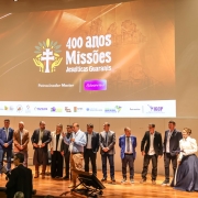 Foto mostra autoridades no palco em fileira. No alto, uma tela mostra uma proje&ccedil;&atilde;o com a logo do evento dos 400 anos das Miss&otilde;es Jesu&iacute;ticas Guaranis.