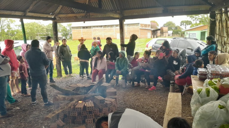 A foto mostra um grupo grande de pessoas reunidas sob um quiosque r&uacute;stico, com estrutura de madeira. No centro do espa&ccedil;o h&aacute; uma fogueira acesa, cercada por tijolos empilhados de maneira circular, com lenhas queimando e fuma&ccedil;a subindo levemente. As pessoas est&atilde;o distribu&iacute;das ao redor da fogueira, algumas sentadas em bancos de madeira e outras em p&eacute; nas laterais do abrigo. &Agrave; direita, sobre uma mesa r&uacute;stica, h&aacute; sacolas pl&aacute;sticas e recipientes com alimentos. 