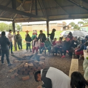 A foto mostra um grupo grande de pessoas reunidas sob um quiosque r&uacute;stico, com estrutura de madeira. No centro do espa&ccedil;o h&aacute; uma fogueira acesa, cercada por tijolos empilhados de maneira circular, com lenhas queimando e fuma&ccedil;a subindo levemente. As pessoas est&atilde;o distribu&iacute;das ao redor da fogueira, algumas sentadas em bancos de madeira e outras em p&eacute; nas laterais do abrigo. &Agrave; direita, sobre uma mesa r&uacute;stica, h&aacute; sacolas pl&aacute;sticas e recipientes com alimentos. 