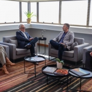 Esta &eacute; uma descri&ccedil;&atilde;o detalhada da imagem para fins de acessibilidade:

Descri&ccedil;&atilde;o da Imagem
A fotografia registra uma reuni&atilde;o institucional em um escrit&oacute;rio moderno, focada no combate &agrave; sonega&ccedil;&atilde;o fiscal.

Participantes: Quatro pessoas (tr&ecirc;s homens e uma mulher) est&atilde;o sentadas em poltronas individuais cinzas, dispostas em formato de "U" para facilitar o di&aacute;logo.

&Agrave; esquerda, uma mulher de cabelos loiros veste uma blusa verde escura e cal&ccedil;as bege.

Ao centro-esquerda, um homem de cabelos grisalhos e &oacute;culos veste um terno azul marinho e gesticula enquanto fala.

Ao centro-direita, outro homem de cabelos grisalhos veste um palet&oacute; cinza claro sobre uma camisa azul.

&Agrave; direita, um homem de cabelos escuros veste um terno preto e observa a conversa.

Ambiente:

O grupo est&aacute; sobre um tapete listrado em tons de marrom, laranja e bege.

&Agrave; frente deles, h&aacute; tr&ecirc;s pequenas mesas de centro circulares pretas; sobre elas, veem-se livros de arte/fotografia e alguns documentos.

Ao fundo, grandes janelas de vidro ocupam toda a extens&atilde;o da parede, revelando uma vista urbana clara, embora os detalhes externos estejam ofuscados pela luminosidade. Um pequeno vaso com uma planta verde est&aacute; posicionado no parapeito da janela.

Contexto: De acordo com o t&iacute;tulo do arquivo, a imagem retrata a entrega de dados pela Receita Estadual ao Minist&eacute;rio P&uacute;blico sobre casos de sonega&ccedil;&atilde;o que superam R$ 217 milh&otilde;es.

Clima: A ilumina&ccedil;&atilde;o &eacute; natural e abundante, conferindo um aspecto s&oacute;brio e profissional ao encontro.