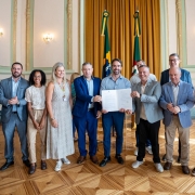 Esta &eacute; uma descri&ccedil;&atilde;o detalhada da imagem para fins de acessibilidade:

Descri&ccedil;&atilde;o da Imagem
A fotografia registra o ato oficial de lan&ccedil;amento do programa Passe F&aacute;cil Estudantil, realizado em um sal&atilde;o solene com arquitetura cl&aacute;ssica.

A&ccedil;&atilde;o Central: No centro da imagem, o Governador Eduardo Leite (vestindo camisa social cinza azulada e cal&ccedil;a escura) e outros dois homens seguram juntos um documento oficial aberto com duas p&aacute;ginas, exibindo-o para a c&acirc;mera.

O Grupo: Ao redor deles, outras sete pessoas (cinco homens e duas mulheres) posam para a foto em uma linha horizontal. O grupo veste trajes que variam entre o social e o casual-executivo. Uma das mulheres &agrave; esquerda utiliza um crach&aacute; oficial com cord&atilde;o colorido.

O Ambiente:

O grupo est&aacute; sobre um piso de madeira (parquet) com padr&otilde;es geom&eacute;tricos claros e escuros.

Ao fundo, h&aacute; um grande painel de cortinas douradas que emoldura as bandeiras do Brasil e do Rio Grande do Sul, posicionadas em mastros centrais.

As paredes s&atilde;o de tom creme com detalhes em relevo verde claro e arandelas cl&aacute;ssicas acesas.

&Agrave; esquerda, nota-se uma caixa de som sobre um pedestal, indicando a realiza&ccedil;&atilde;o de uma cerim&ocirc;nia com sistema de &aacute;udio.

Contexto: Conforme o t&iacute;tulo do arquivo, o evento marca uma iniciativa para ampliar o acesso &agrave; educa&ccedil;&atilde;o no estado atrav&eacute;s do Passe F&aacute;cil Estudantil.