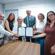 Esta &eacute; uma descri&ccedil;&atilde;o detalhada da imagem para fins de acessibilidade:

Descri&ccedil;&atilde;o da Imagem
A fotografia registra o ato formal de assinatura de um conv&ecirc;nio em um ambiente de escrit&oacute;rio com ilumina&ccedil;&atilde;o clara e paredes brancas.

A&ccedil;&atilde;o Central: Um grupo de seis pessoas (quatro mulheres e dois homens) est&aacute; posicionado atr&aacute;s de uma mesa de madeira clara. Eles seguram juntos uma pasta oficial preta aberta, exibindo um documento de duas p&aacute;ginas com o bras&atilde;o do Estado do Rio Grande do Sul no topo.

Os Participantes:

&Agrave; esquerda, um homem de camisa social clara e &oacute;culos sorri para a c&acirc;mera.

Ao lado dele, uma mulher de blusa preta sem mangas.

No centro, uma mulher de cabelos loiros curtos, usando &oacute;culos e um crach&aacute; oficial com cord&atilde;o colorido, segura a pasta.

Atr&aacute;s dela, um homem de terno cinza e gravata azul tamb&eacute;m segura o documento.

&Agrave; direita, uma mulher de blusa branca e outra de vestido marrom completam o grupo. Todos sorriem, transmitindo um clima de realiza&ccedil;&atilde;o.

Detalhes do Ambiente:

Sobre a mesa, em frente ao grupo, h&aacute; apenas uma caneta azul, indicando que a assinatura acabou de ocorrer.

O fundo &eacute; simples, composto por divis&oacute;rias de escrit&oacute;rio brancas, o que mant&eacute;m o foco total nas pessoas e no documento.

Contexto: De acordo com o t&iacute;tulo do arquivo, a imagem celebra a habilita&ccedil;&atilde;o do munic&iacute;pio de Torres ao programa TEAcolhe, com validade para o ano de 2026.