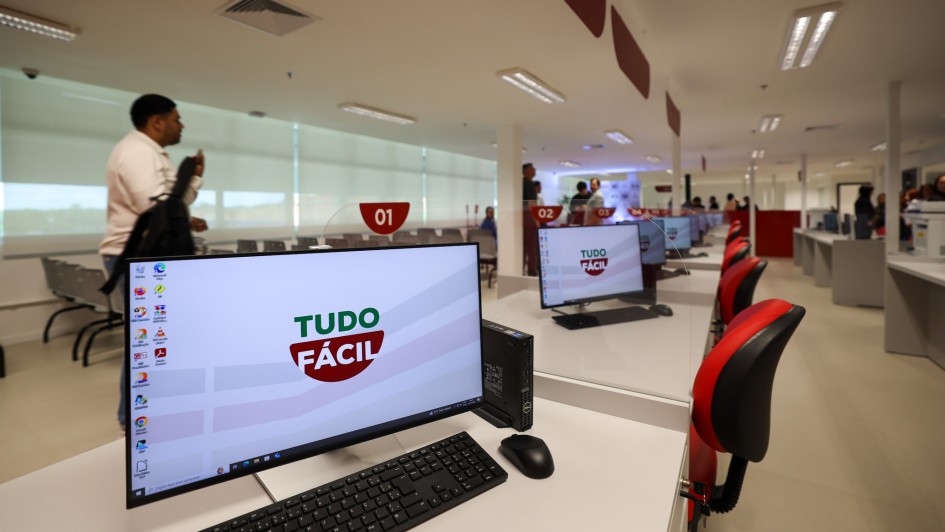 Foto de uma unidade do Tudo F&aacute;cil, de paredes e piso branco, com foco numa mesa de atendimento com computador. H&aacute; v&aacute;rias outras alinhadas atr&aacute;s. H&aacute; pessoas andando pelo local.