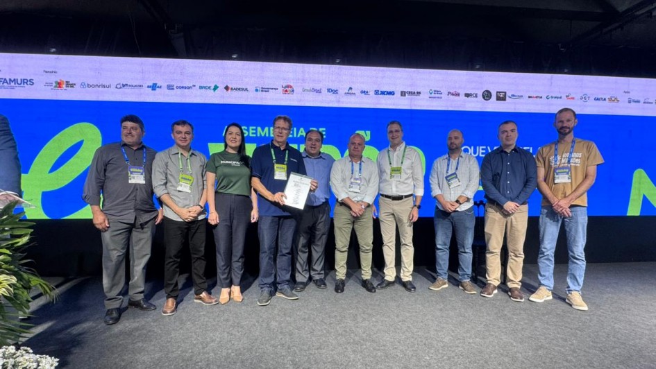 Foto posada de dez pessoas de p&eacute;, enfileiradas, de frente para c&acirc;mera. Elas se encontram num ambiente fechado, com um tel&atilde;o ao fundo. Uma delas exibe um papel para a c&acirc;mera.