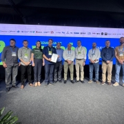 Foto posada de dez pessoas de p&eacute;, enfileiradas, de frente para c&acirc;mera. Elas se encontram num ambiente fechado, com um tel&atilde;o ao fundo. Uma delas exibe um papel para a c&acirc;mera.