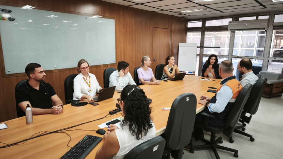 Esta &eacute; uma descri&ccedil;&atilde;o detalhada da &uacute;ltima imagem para fins de acessibilidade:

Descri&ccedil;&atilde;o da Imagem
A fotografia registra uma reuni&atilde;o de trabalho t&eacute;cnica realizada em uma sala de reuni&otilde;es de estilo contempor&acirc;neo e funcional.

A&ccedil;&atilde;o Central: Um grupo de nove pessoas (cinco mulheres e quatro homens) est&aacute; reunido em torno de uma grande mesa de madeira clara em formato oval.

Os Participantes:

&Agrave; esquerda, tr&ecirc;s participantes est&atilde;o sentados, incluindo uma mulher de camisa branca e &oacute;culos vermelhos que utiliza um notebook.

Ao fundo, na cabeceira oposta, uma mulher gesticula com as m&atilde;os enquanto fala para o grupo.

&Agrave; direita, em destaque, um homem veste um colete da Defesa Civil (azul marinho com a parte superior em laranja e o tri&acirc;ngulo s&iacute;mbolo da institui&ccedil;&atilde;o nas costas), indicando o car&aacute;ter operacional e de gest&atilde;o de riscos da reuni&atilde;o.

O Ambiente:

A sala possui paredes revestidas com pain&eacute;is de madeira.

H&aacute; um grande quadro branco (ou tela de vidro) na parede lateral esquerda, que exibe o logotipo da Secretaria de Inova&ccedil;&atilde;o, Ci&ecirc;ncia e Tecnologia do Rio Grande do Sul.

Ao fundo, grandes janelas de vidro mostram uma vista urbana, e um quadro branco tipo flip chart est&aacute; posicionado no canto.

O teto &eacute; composto por placas modulares com lumin&aacute;rias embutidas que garantem uma ilumina&ccedil;&atilde;o clara e uniforme.

Contexto: Segundo o t&iacute;tulo do arquivo, trata-se de uma reuni&atilde;o do Conselho Consultivo do Criec (Centro de Resili&ecirc;ncia e Intelig&ecirc;ncia Estrat&eacute;gica para o Cuidado), focada em consolidar a governan&ccedil;a para o enfrentamento de desafios clim&aacute;ticos no estado.