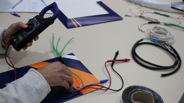 Foto em plano detalhe das m&atilde;os de um socioeducando mexendo com um aparelho eletr&ocirc;nico, com os bra&ccedil;os apoiados numa mesa clara. Sobre ela est&atilde;o fios e papeis. 