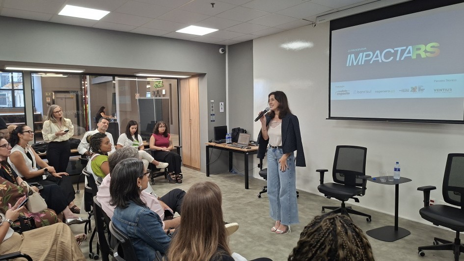 Foto mostra palestrante diante de plateia e tel&atilde;o ao fundo com o nome do Programa Impacta RS.