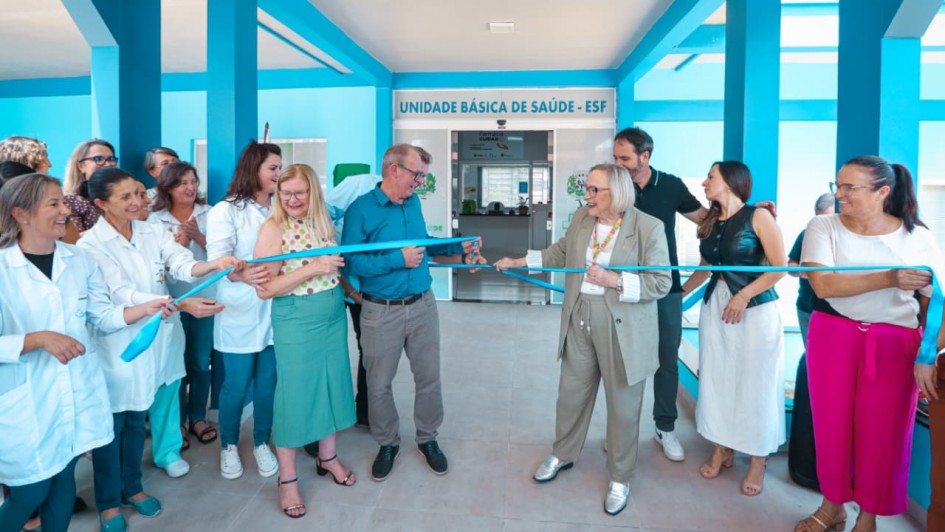 A foto mostra um grupo de pessoas reunidas na entrada de uma Unidade B&aacute;sica de Sa&uacute;de &ndash; ESF, identificada por uma placa localizada acima da porta principal. O pr&eacute;dio tem estrutura em tons de azul vibrante. No centro da imagem, uma faixa azul est&aacute; sendo cortada em uma cerim&ocirc;nia de inaugura&ccedil;&atilde;o, segurada por v&aacute;rias pessoas alinhadas dos dois lados. 