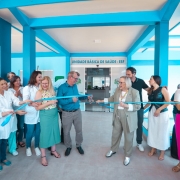 A foto mostra um grupo de pessoas reunidas na entrada de uma Unidade B&aacute;sica de Sa&uacute;de &ndash; ESF, identificada por uma placa localizada acima da porta principal. O pr&eacute;dio tem estrutura em tons de azul vibrante. No centro da imagem, uma faixa azul est&aacute; sendo cortada em uma cerim&ocirc;nia de inaugura&ccedil;&atilde;o, segurada por v&aacute;rias pessoas alinhadas dos dois lados. 