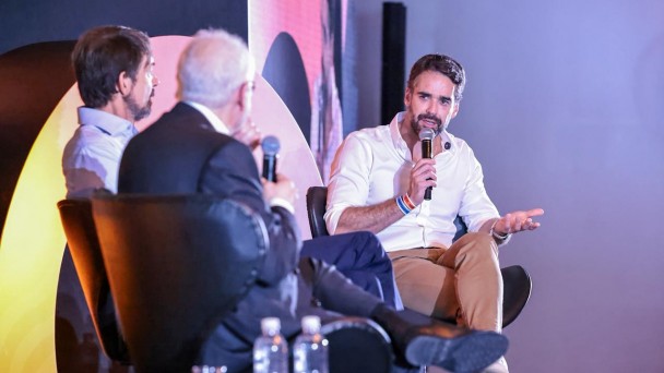 Tr&ecirc;s homens sentados em poltronas participam de uma conversa em um palco iluminado; um deles fala ao microfone enquanto os outros escutam atentamente.