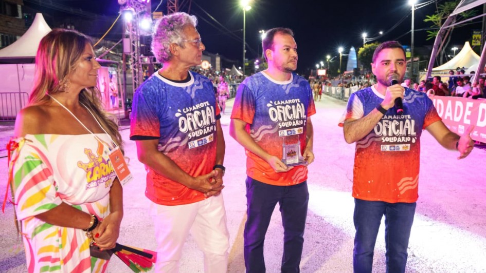 Quatro pessoas est&atilde;o em uma avenida iluminada &agrave; noite durante um evento festivo; tr&ecirc;s homens vestem camisetas coloridas com o texto &ldquo;Camarote Oficial&rdquo; e uma mulher usa um vestido estampado, enquanto um dos homens fala ao microfone.