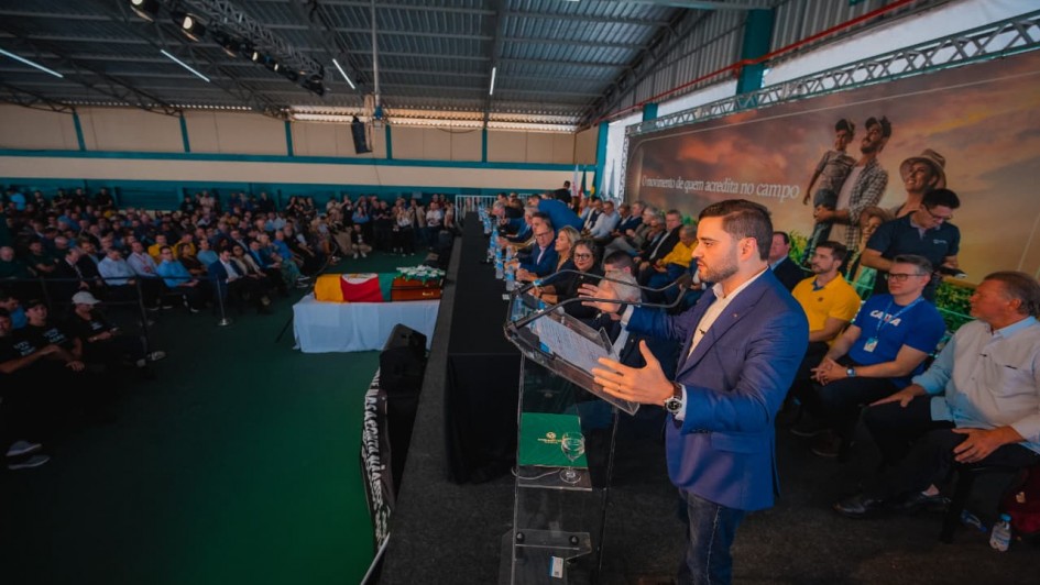 Evento em um pavilh&atilde;o amplo, com palco elevado onde v&aacute;rias pessoas est&atilde;o sentadas ao longo de uma mesa. Em destaque, o vice-governador fala ao microfone em um p&uacute;lpito transparente, enquanto o p&uacute;blico ocupa cadeiras ao fundo.