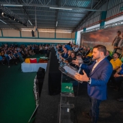 Evento em um pavilh&atilde;o amplo, com palco elevado onde v&aacute;rias pessoas est&atilde;o sentadas ao longo de uma mesa. Em destaque, o vice-governador fala ao microfone em um p&uacute;lpito transparente, enquanto o p&uacute;blico ocupa cadeiras ao fundo.