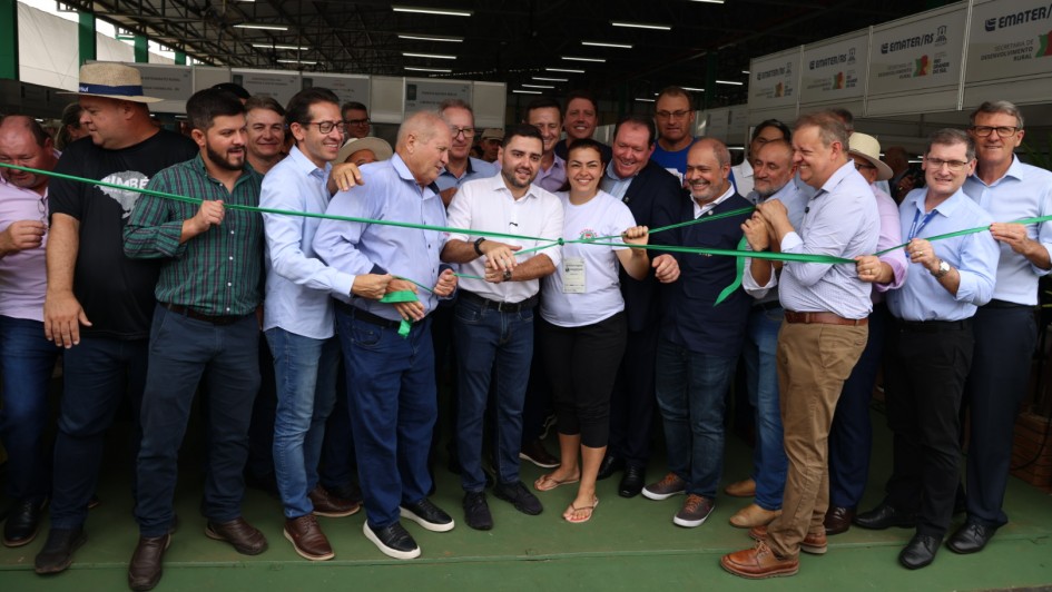 Esta &eacute; uma descri&ccedil;&atilde;o detalhada da imagem para fins de acessibilidade:

Descri&ccedil;&atilde;o da Imagem
A fotografia registra a cerim&ocirc;nia de inaugura&ccedil;&atilde;o do Pavilh&atilde;o da Agricultura Familiar durante a feira Expodireto Cotrijal.

A&ccedil;&atilde;o Central: No centro da imagem, um grande grupo de homens e uma mulher est&atilde;o reunidos para o ato simb&oacute;lico de corte da fita inaugural. A fita &eacute; de cor verde vibrante. V&aacute;rias m&atilde;os seguram e tensionam a fita enquanto o Governador Eduardo Leite (ao centro, de camisa branca e cal&ccedil;a jeans) se prepara para cort&aacute;-la.

Os Participantes:

O grupo &eacute; composto por cerca de 20 pessoas, a maioria homens vestindo camisas sociais de tons claros (azul e branco) ou camisetas de eventos.

&Agrave; direita do Governador, uma mulher de camiseta branca e cabelos presos sorri enquanto segura a fita.

Os participantes apresentam express&otilde;es de entusiasmo e alegria, celebrando o momento.

O Ambiente:

A cena ocorre sob a estrutura de um pavilh&atilde;o com teto de metal aparente e ilumina&ccedil;&atilde;o por calhas de LED.

Ao fundo, &agrave; direita, veem-se estandes brancos com logotipos da EMATER/RS e da Secretaria de Desenvolvimento Rural.

O ch&atilde;o do pavilh&atilde;o &eacute; pintado de verde escuro.

Contexto: Segundo o t&iacute;tulo do arquivo, o espa&ccedil;o destaca a diversidade da produ&ccedil;&atilde;o rural do Rio Grande do Sul no contexto da feira Expodireto Cotrijal.