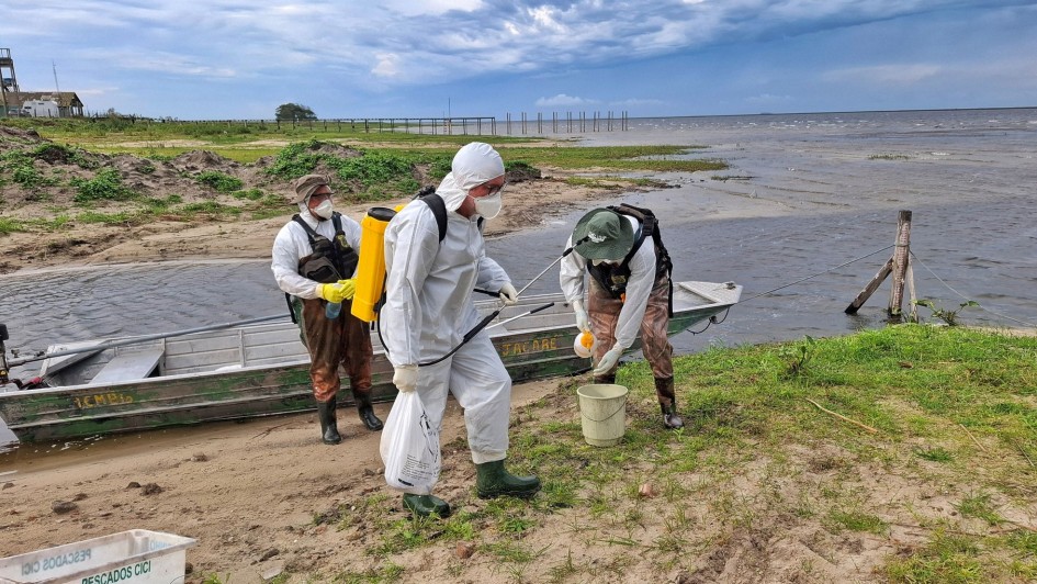 Foto mostra tr&ecirc;s servidores com roupas brancas, botas e m&aacute;scaras, preparando produtos para aplica&ccedil;&atilde;o. Eles est&atilde;o na beira da lagoa, pr&oacute;ximo a um barco. Ao fundo, a lagoa e o c&eacute;u carregado para chuva. 