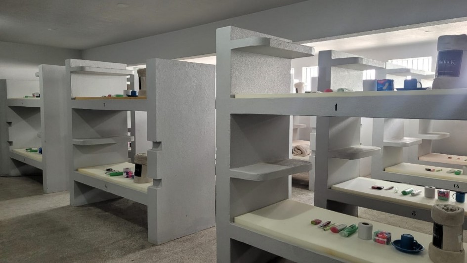 A foto mostra um ambiente interno com fileiras de estruturas cinzas que funcionam como beliches ou alojamentos modulares, cada uma com prateleiras embutidas. Sobre as superf&iacute;cies est&atilde;o organizados itens pessoais, como produtos de higiene e cobertores.