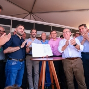 Esta &eacute; uma descri&ccedil;&atilde;o da imagem para fins de acessibilidade:

A fotografia registra um momento de celebra&ccedil;&atilde;o oficial durante a Expodireto Cotrijal, em que um grupo de autoridades e representantes do setor agropecu&aacute;rio se re&uacute;ne para a assinatura de documentos de investimento.

Elementos Principais
O Ato Central: Ao centro da imagem, sobre uma mesa alta e redonda de madeira (estilo bistr&ocirc;), um documento oficial aberto &eacute; exibido para a c&acirc;mera. V&aacute;rias m&atilde;os o seguram levemente pelas bordas.

As Autoridades (da esquerda para a direita):

Em destaque &agrave; esquerda, o vice-governador Gabriel Souza, vestindo uma camisa social azul-marinho e cal&ccedil;a jeans, aplaude com um sorriso.

Logo atr&aacute;s dele, ligeiramente ao centro, o governador Eduardo Leite, vestindo uma camisa azul clara de mangas curtas, observa o documento com uma express&atilde;o sorridente.

&Agrave; direita da mesa, um homem de camisa branca e cal&ccedil;a c&aacute;qui aplaude com entusiasmo.

Diversos outros homens em trajes casuais e sociais comp&otilde;em o grupo ao fundo, todos sorrindo ou aplaudindo, refor&ccedil;ando o clima de conquista.

Detalhes e Ambiente
Cen&aacute;rio: O evento ocorre sob uma grande tenda branca, com estruturas met&aacute;licas vis&iacute;veis no teto. Ao fundo, divis&oacute;rias de stands indicam que o local &eacute; um pavilh&atilde;o de feiras.

Ilumina&ccedil;&atilde;o e Composi&ccedil;&atilde;o: A foto foi tirada de um &acirc;ngulo ligeiramente de baixo para cima (contra-picado), o que confere uma sensa&ccedil;&atilde;o de import&acirc;ncia ao momento. A luz &eacute; clara e bem distribu&iacute;da, t&iacute;pica de eventos diurnos em ambientes cobertos.

Clima: A atmosfera &eacute; de otimismo e coopera&ccedil;&atilde;o institucional, evidenciada pela linguagem corporal de celebra&ccedil;&atilde;o (palmas e sorrisos) de todos os presentes.